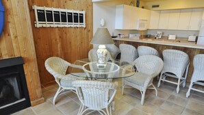 Un coin repas avec des chaises en osier et une table en verre, une cheminée, et une cuisine en arrière-plan.