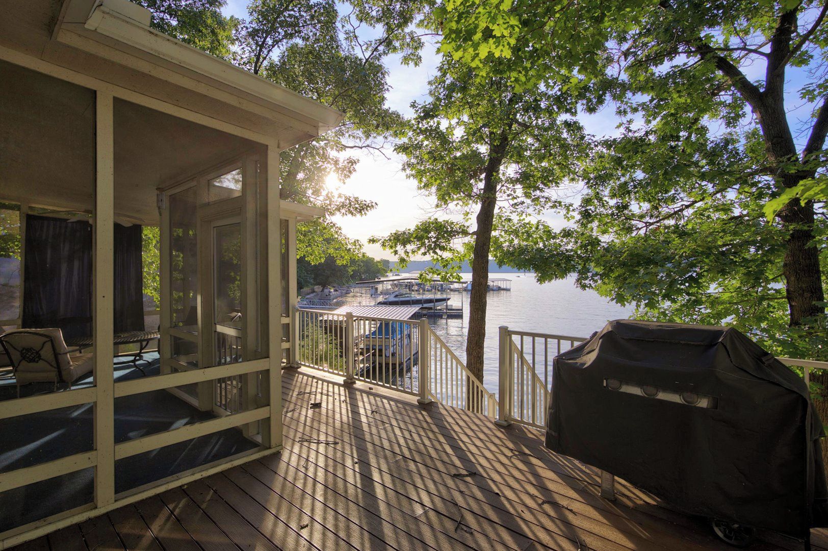 A covered deck with a grill, chairs, and a view of boats on a lake.