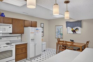 Una cocina moderna con armarios de madera, un refrigerador blanco, un microondas y una estufa. Hay una mesa de comedor con sillas y una ventana con un gallo decorativo.