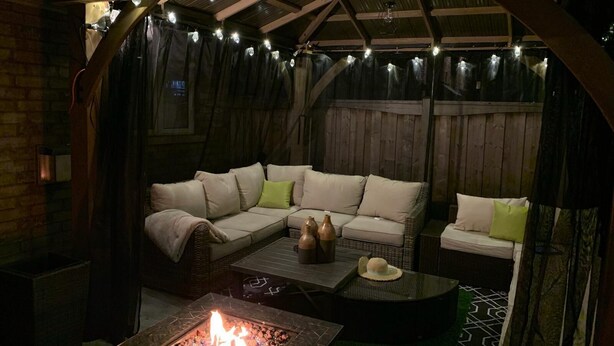 Un espacio exterior con asientos al aire libre, una chimenea, sofás acolchados e iluminación con luces colgantes.