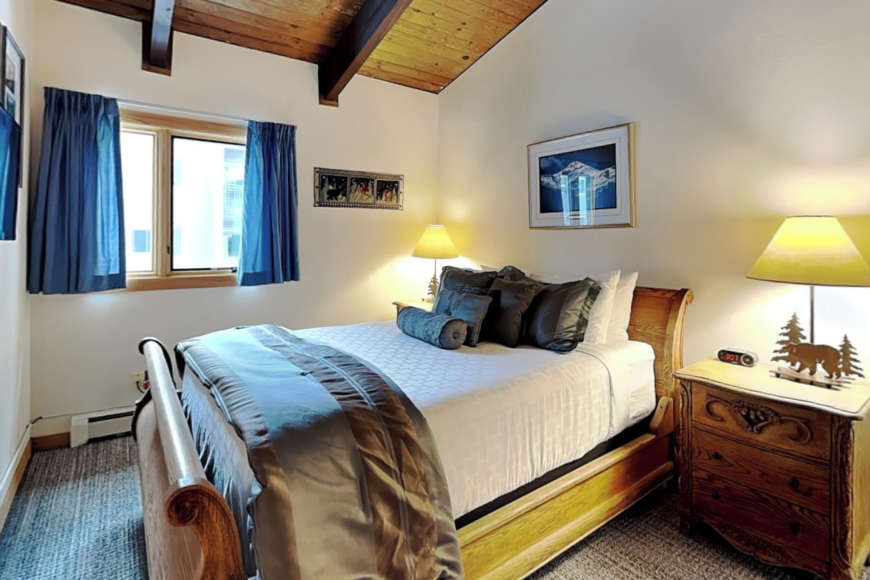 A bedroom with a bed, a nightstand, a lamp, a framed picture, and a window with curtains.