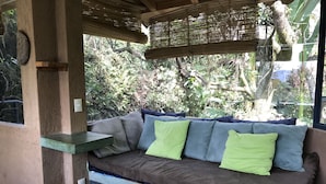 A cozy outdoor seating area with a cushioned bench, a small shelf, and a view of lush greenery.