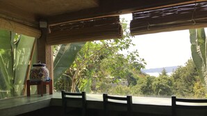A room with a large window overlooking a lush garden and a distant view of hills. There are two wooden chairs and a decorative ceramic pot on a small table.