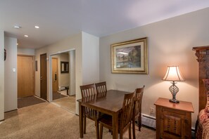 A hotel room with a wooden dining table, chairs, a small cabinet, and a lamp. There is a framed picture on the wall and a door leading to another room.