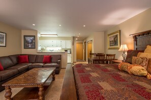 A living room with a brown sectional sofa, a wooden coffee table, and a patterned bedspread. There is a kitchen in the background with white cabinets and a dining area with wooden chairs.
