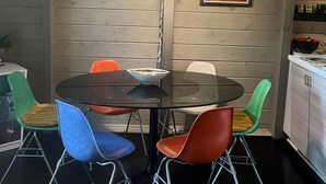 Een eetruimte met een ronde tafel, kleurrijke stoelen, een hanglamp en een schilderij aan de muur.