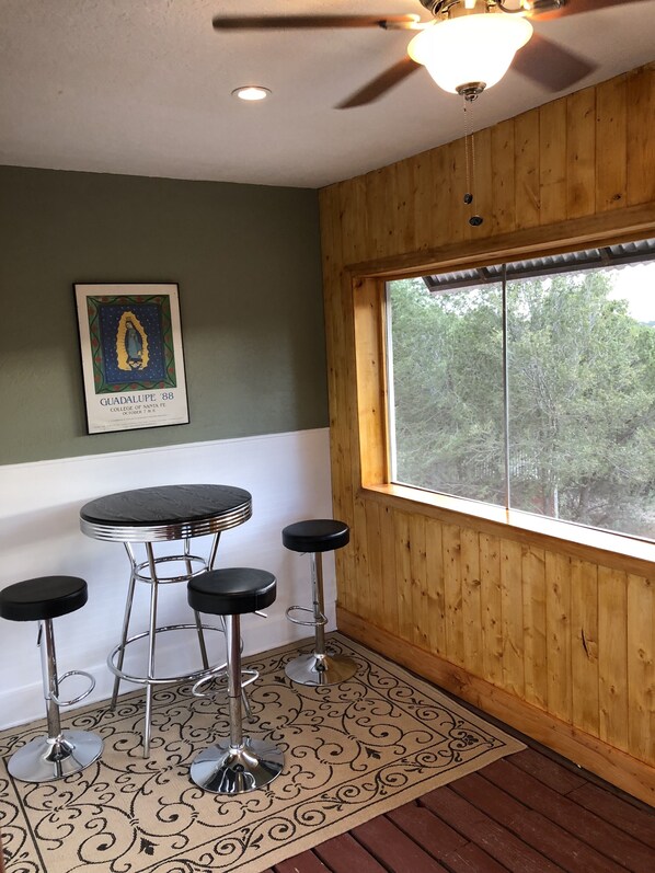 A room with a wooden paneled wall, a ceiling fan, a framed picture, a round table with four bar stools, and a large window with a view of trees.