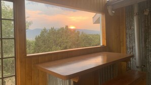 A room with a large window overlooking a forested area and mountains during sunset.
