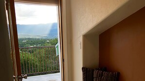 A room with a wooden floor, a sliding glass door leading to a balcony with a view of greenery and distant mountains.