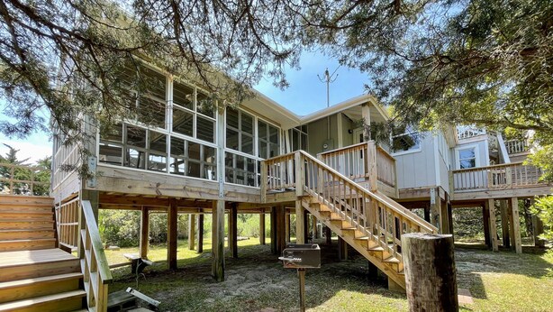 A wooden deck with a staircase leading to a second level, surrounded by trees. - Kith And Kin:  Old Ocracoke style cottage, spacious screened porch. (Ocracoke)