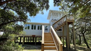 A wooden deck with a staircase leading to a house elevated on stilts, surrounded by trees. - Kith And Kin:  Old Ocracoke style cottage, spacious screened porch. (Ocracoke)
