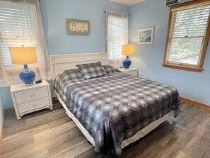 A bedroom with a bed, two bedside tables with lamps, a nightstand, a window with blinds, and a wall-mounted picture.