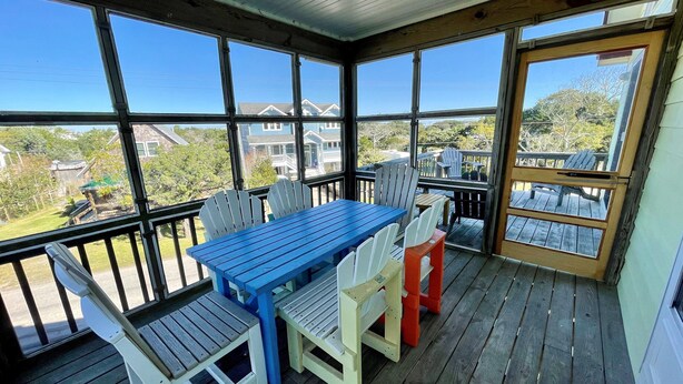 Ein überdachter Außenbereich mit blauem Tisch und Stühlen, einer Holzterrasse und Blick auf Häuser und Bäume.