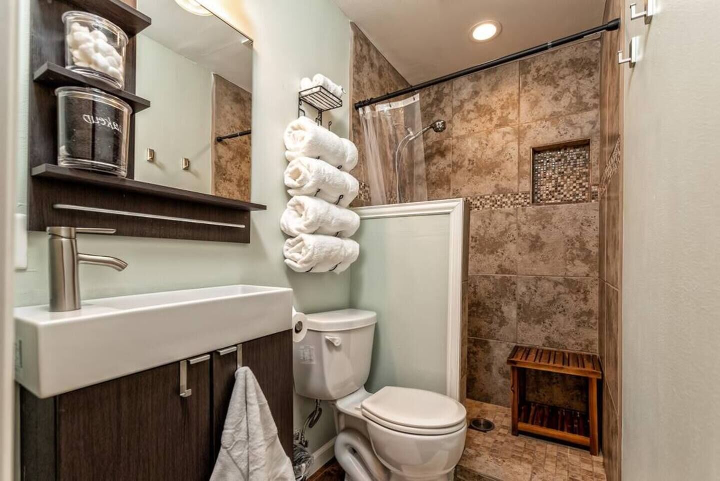 A modern bathroom with a sink, toilet, and shower. There are towels hanging on a rack and a small shelf with containers.