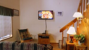 Ein gemütliches Wohnzimmer mit einem karierten Sofa, einem Holztisch, einem an der Wand montierten Fernseher und einer Treppe mit Holzbalkon.