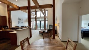 Un vaste espace de vie comprenant un coin repas avec une table et des chaises en bois, une cuisine avec un bar et de grandes fenêtres offrant une vue sur l’extérieur.
