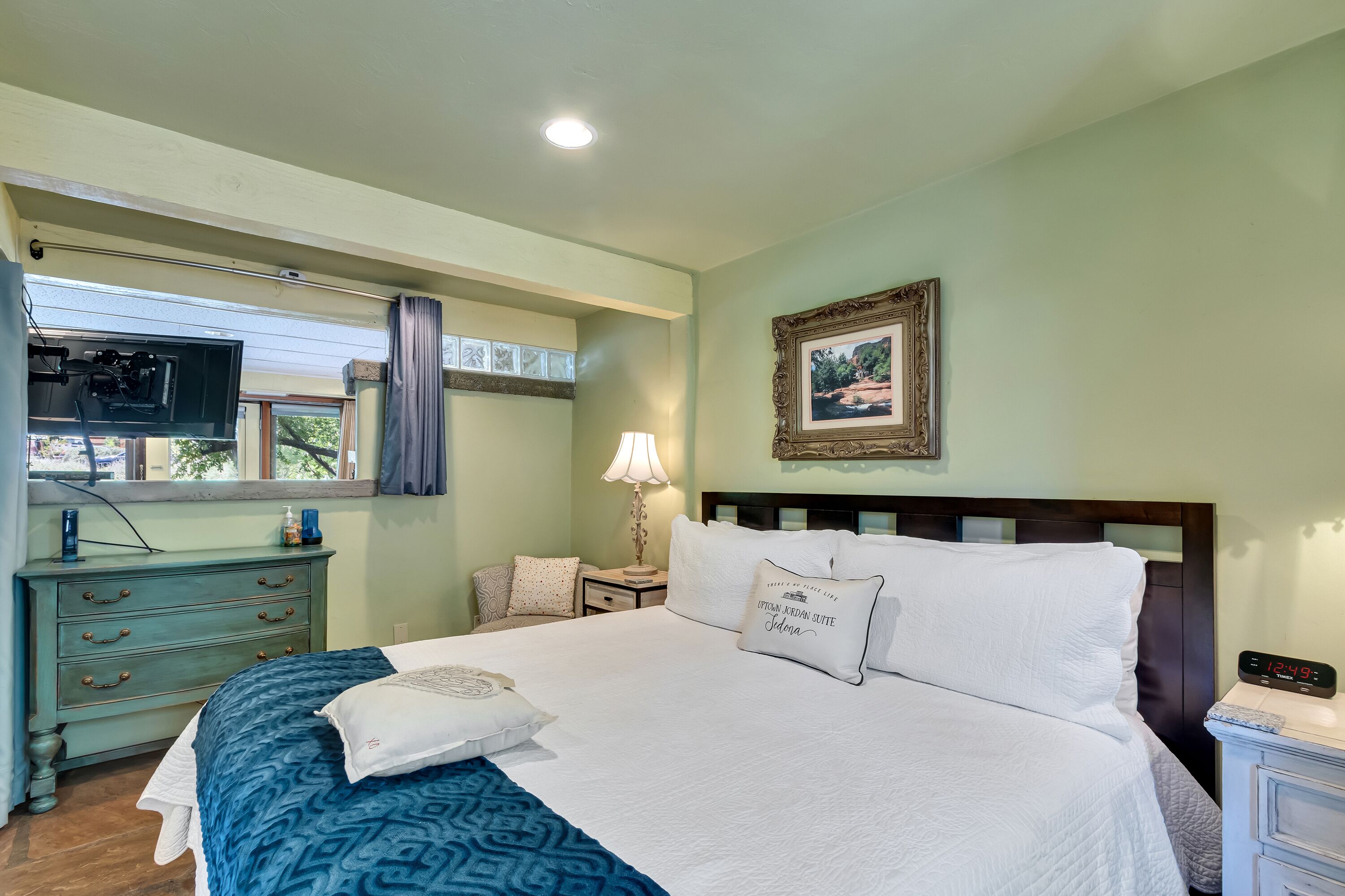 A neatly made bed with white and blue bedding, a bedside table with a lamp, a clock, and a framed picture on the wall.