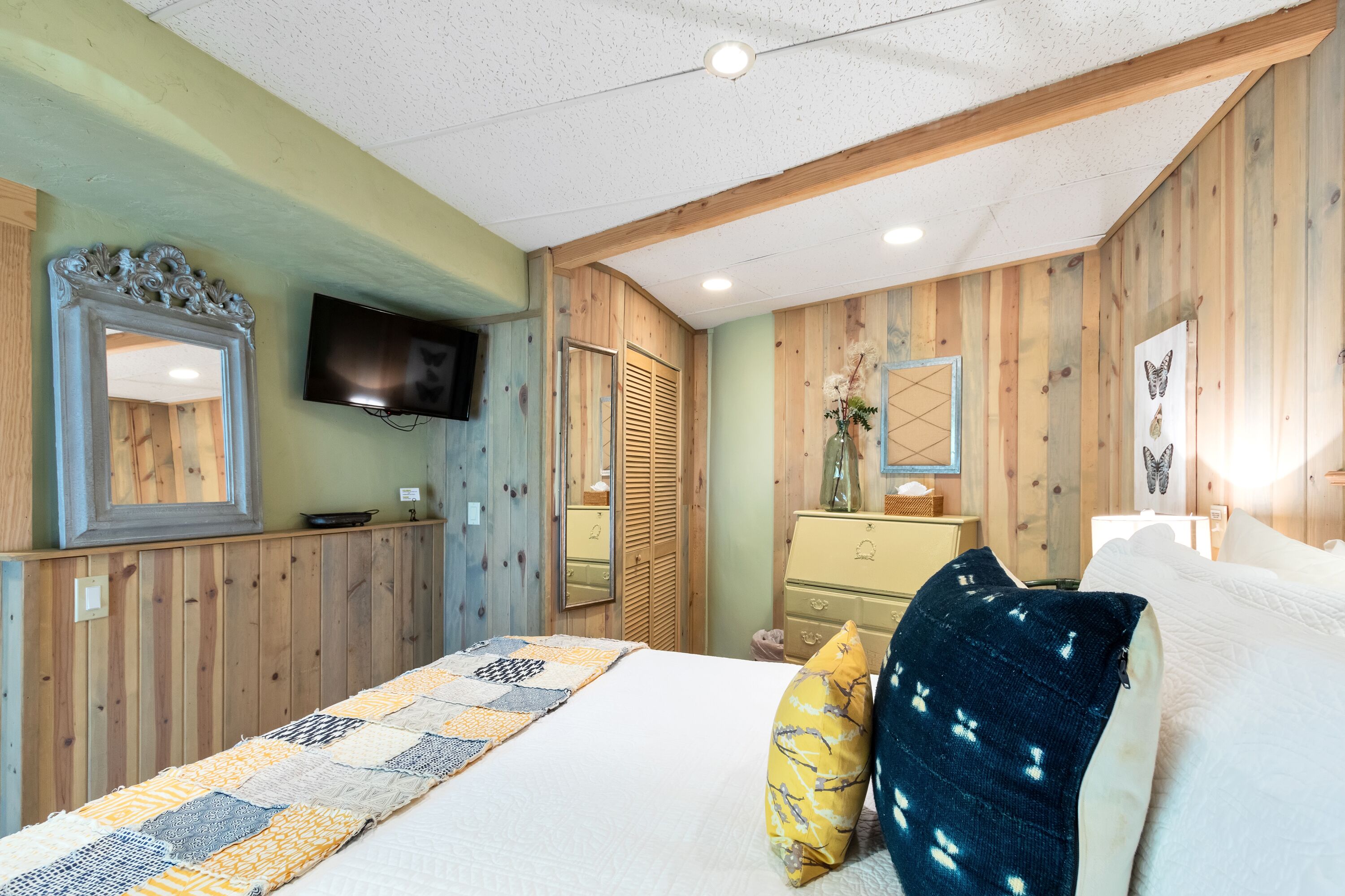 Een slaapkamer met een wand van houten panelen, een bed met quilt, een kapstok, een spiegel en een flatscreen televisie bovenop de kapstok.