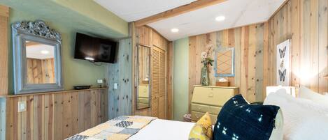 A bedroom with a wooden paneled wall, a bed with a quilt, a dresser, a mirror, and a flat-screen TV mounted above the dresser.