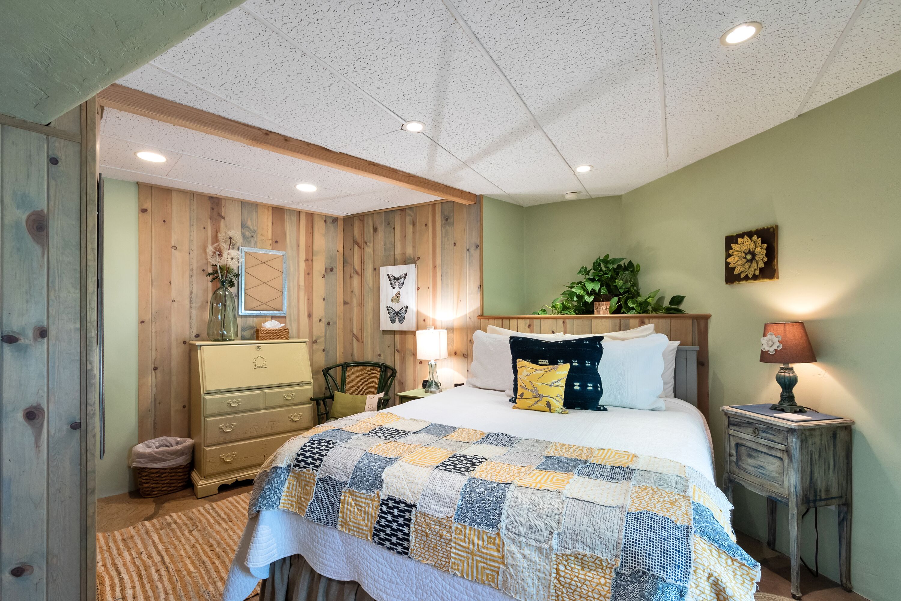 Un dormitorio con cabecera de madera, colcha acolchada, mesita de noche, lámpara y una planta pequeña.