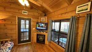 Ein gemütliches Blockhauszimmer mit Holzwänden, einem Kamin, einem Fernseher und einem großen Fenster mit Vorhängen.