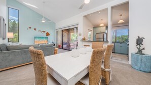 Una sala de estar moderna con un sofá gris, una mesa de comedor blanca, sillas de mimbre y una cocina con vistas al exterior.