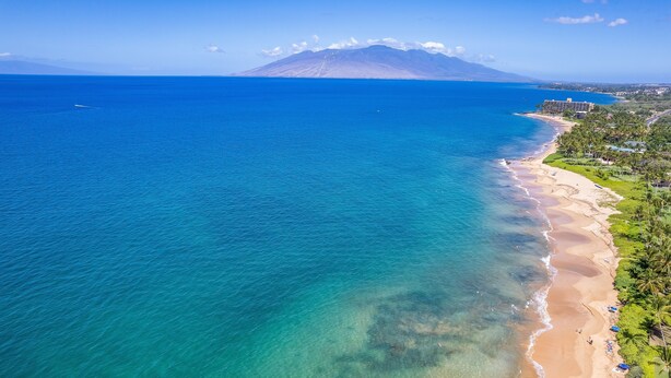 Vista aérea de una zona costera con playa, aguas azul turquesa y una montaña al fondo.