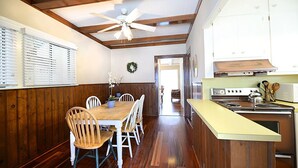 Un coin repas avec une table et des chaises en bois, un ventilateur de plafond, et une cuisine équipée d’une cuisinière et de placards.