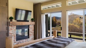 Une chambre avec une cheminée, un téléviseur fixé au-dessus, une grande fenêtre donnant sur l’extérieur et un balcon avec une balustrade en bois.