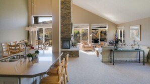 Un vaste espace de vie agrémenté d’une cheminée en pierre, un coin repas avec des chaises en bois et un canapé dans une pièce lumineuse dotée de grandes fenêtres.