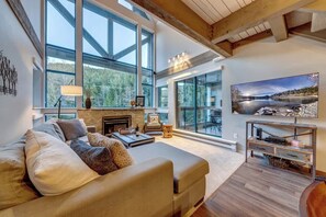 A spacious living room with a large window, a fireplace, a sofa, and a painting. - Greystone Lodge 412 Ski-in/Out by Acer Vacations (Whistler)