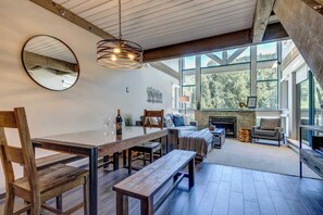 A spacious room with a large dining table, wooden chairs, a fireplace, and large windows offering a view of the outdoors.