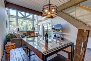 A modern loft with a glass dining table, wooden beams, large windows, and a view of the outdoors.