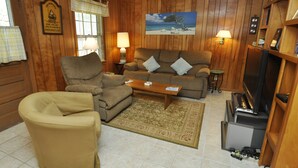 Un salón acogedor con sofá, sillón y mesa de centro, paredes de madera y un cuadro de una playa.