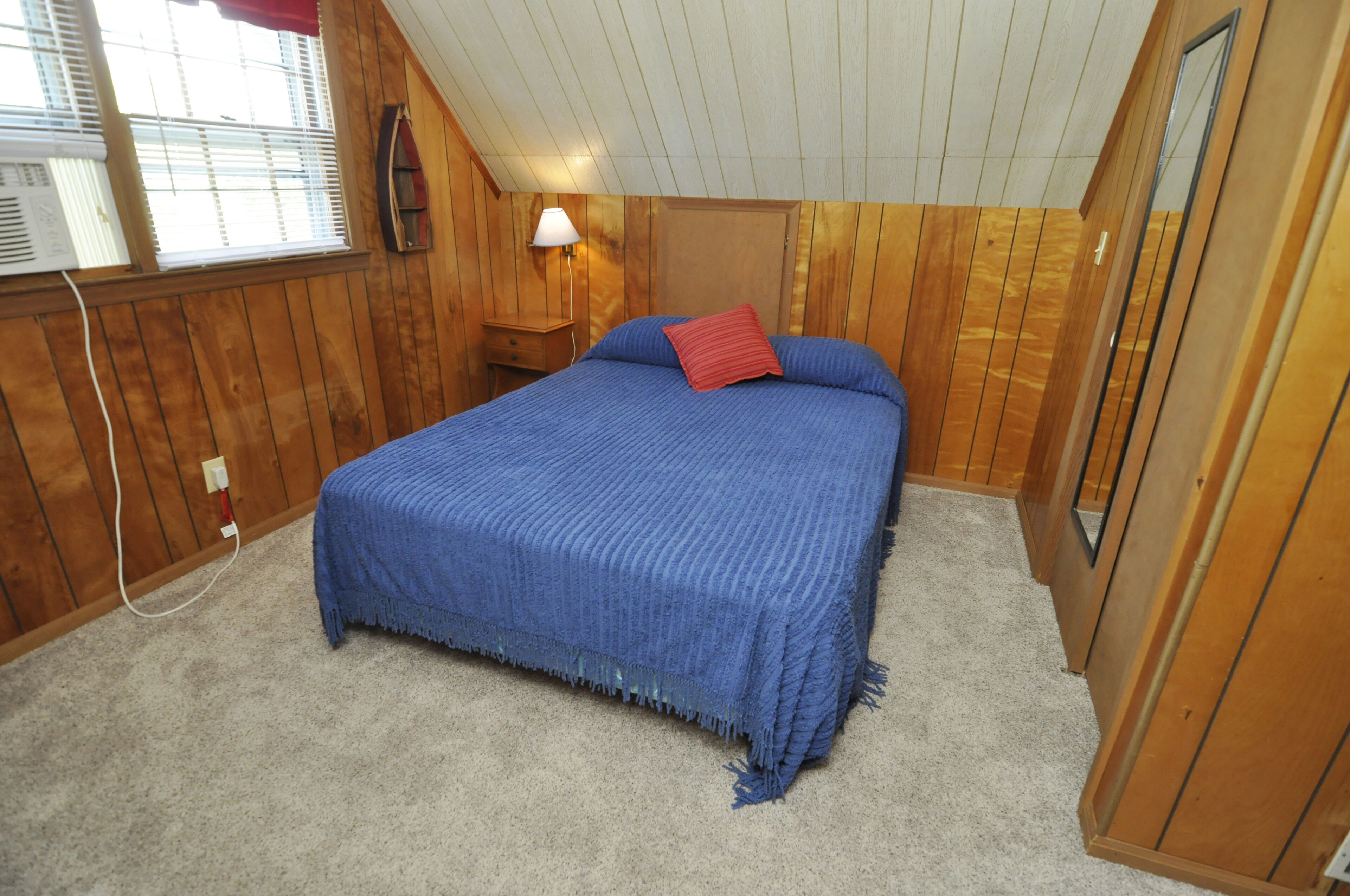A bedroom with a wooden paneled ceiling, a bed with blue bedding, a red pillow, a nightstand with a lamp, and a window with blinds.