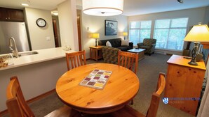 A modern kitchen with a sink, refrigerator, and dining area with a wooden table and chairs. There is a living room with a couch, a lamp, and a window with blinds. - Renovated Ground Floor Condo. Spacious Floor Plan, Pool & Hot Tub Onsite (Whistler)