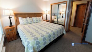 A bedroom with a large bed, two bedside lamps, a nightstand, a full-length mirror, and a door. - Renovated Ground Floor Condo. Spacious Floor Plan, Pool & Hot Tub Onsite (Whistler)
