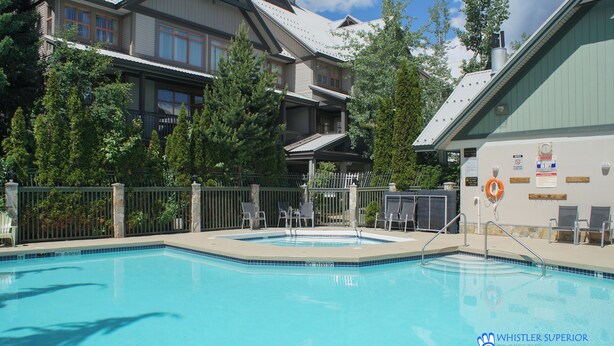 A swimming pool with a fenced deck area, lounge chairs, and a building with multiple balconies in the background. - Renovated Ground Floor Condo. Spacious Floor Plan, Pool & Hot Tub Onsite (Whistler)