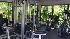 Ein Fitnessraum mit unterschiedlichen Trainingsgeräten, einem Deckenventilator und großen Fenstern mit Blick auf Grünflächen.