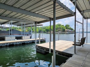 A covered dock with a swing chair, a boat, and a view of a lake and trees.