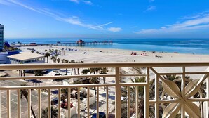 A beach view from a balcony with a clear sky, a pier extending into the ocean, and a sandy beach with umbrellas and palm trees.