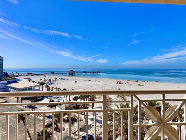 Blick auf einen Strand vom Balkon aus, mit blauem Himmel, einem Steg, der sich ins Meer erstreckt, und einem Sandstrand mit Sonnenschirmen und Palmen.