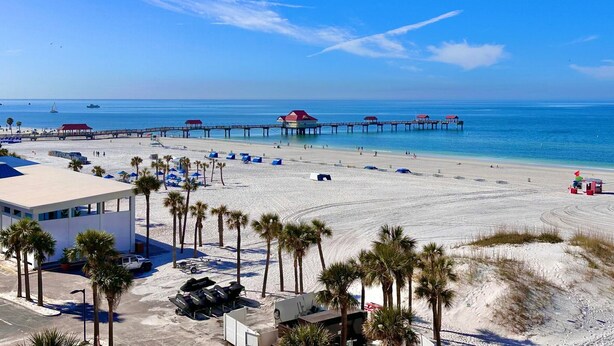 A beach with a pier, palm trees, and beach umbrellas.