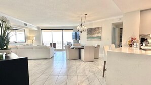 A spacious living area with a large sofa, a dining table with chairs, a chandelier, and a view through large windows.