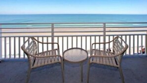 A balcony with two wicker chairs and a small round table, overlooking a beach with a clear view of the ocean.