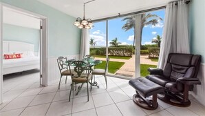 Une chambre avec un lit, une table à manger et des chaises, un fauteuil de massage, et une vue sur des palmiers et un jardin.