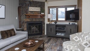 A cozy living room with a stone fireplace, a television, and a comfortable sofa. - Bright, Modern On-Mountain Condo (Whitefish)