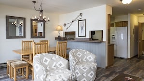 A dining area with a wooden table and chairs, a chandelier, and a kitchen with a refrigerator and cabinets. - Bright, Modern On-Mountain Condo (Whitefish)