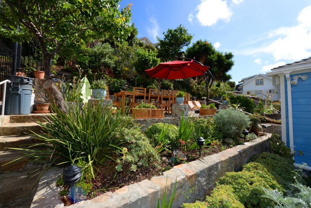 A garden with a wooden bench and a red umbrella, surrounded by various plants and a stone wall.