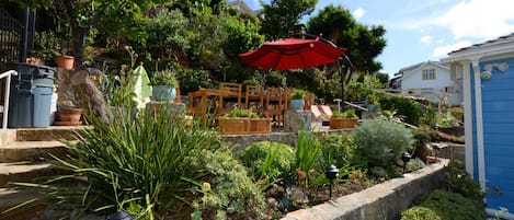 A garden with a wooden bench and a red umbrella, surrounded by various plants and a stone wall.